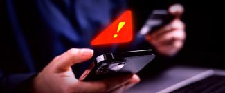 A person holding a smartphone and credit card with a glowing red warning symbol above the phone.